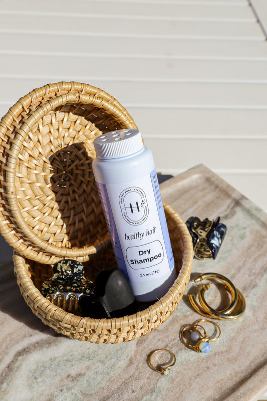 A bottle of dry shampoo with a label that reads 'Dry Shampoo - With Moonstone Crystal'. The bottle is placed inside a woven basket on a wooden surface, alongside some jewelry and decorative items.