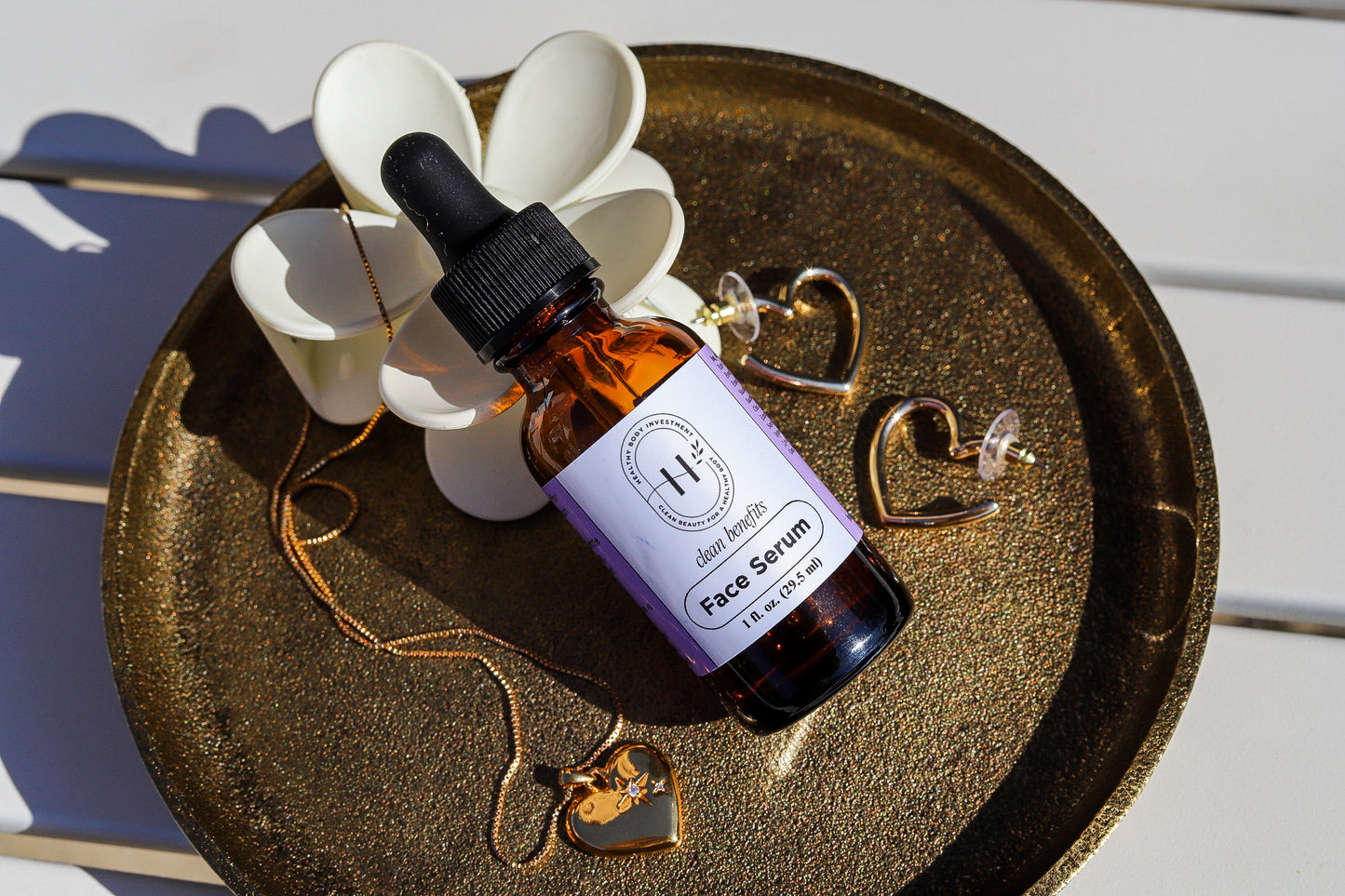 A glass bottle with a dropper containing face serum, placed on a bronze plate next to a white flower and a crystal. The bottle label reads 'FACIAL SERUM'.