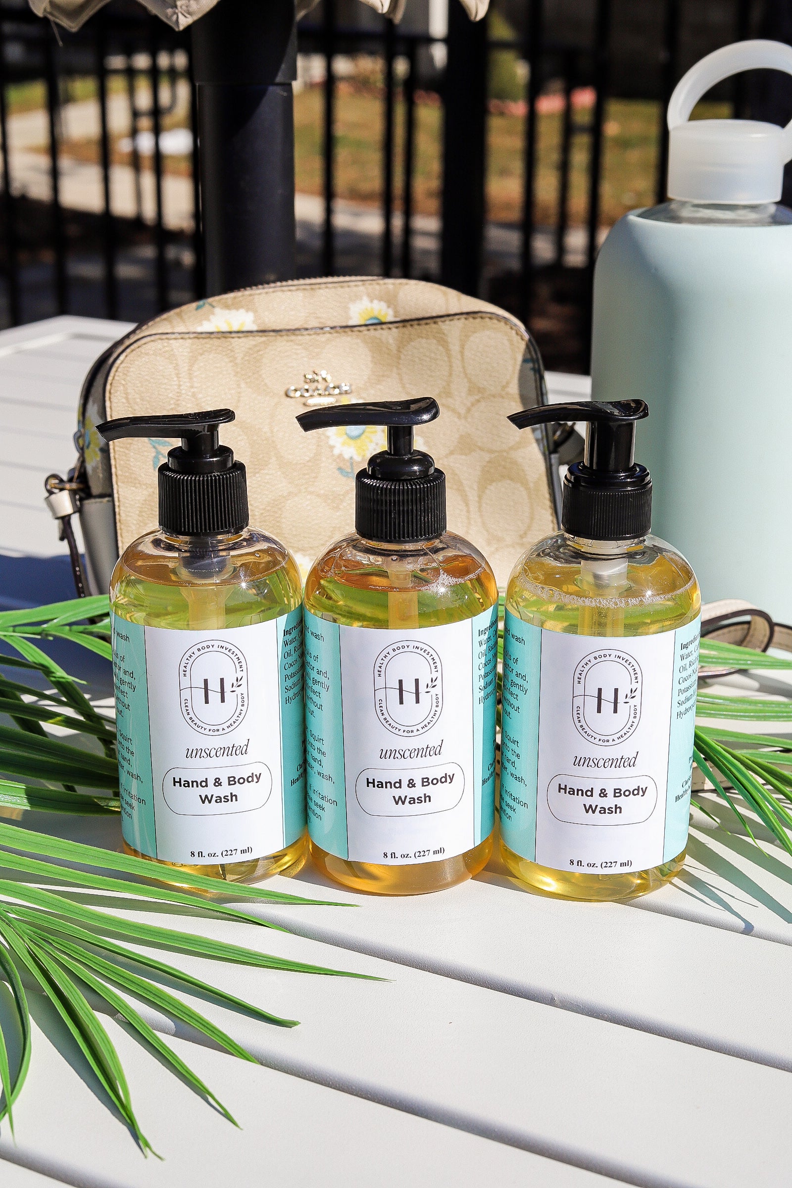Three pump bottles of hand and body soap placed on a surface with a palm leaf in the foreground and a patterned bag in the background.