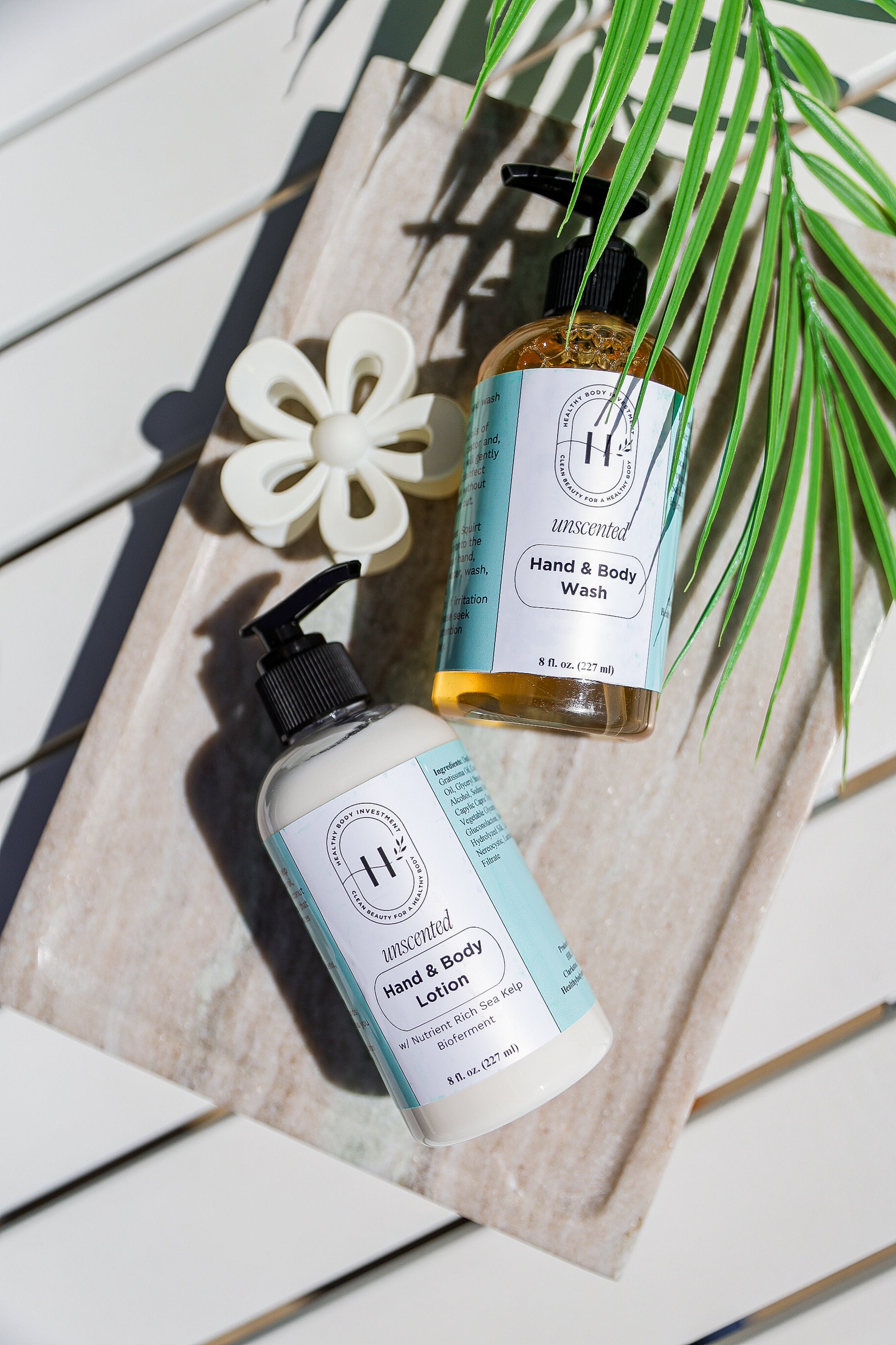 A bottle of hand and body lotion and a bottle of hand and body wash set on a wooden surface with a palm leaf in the background.