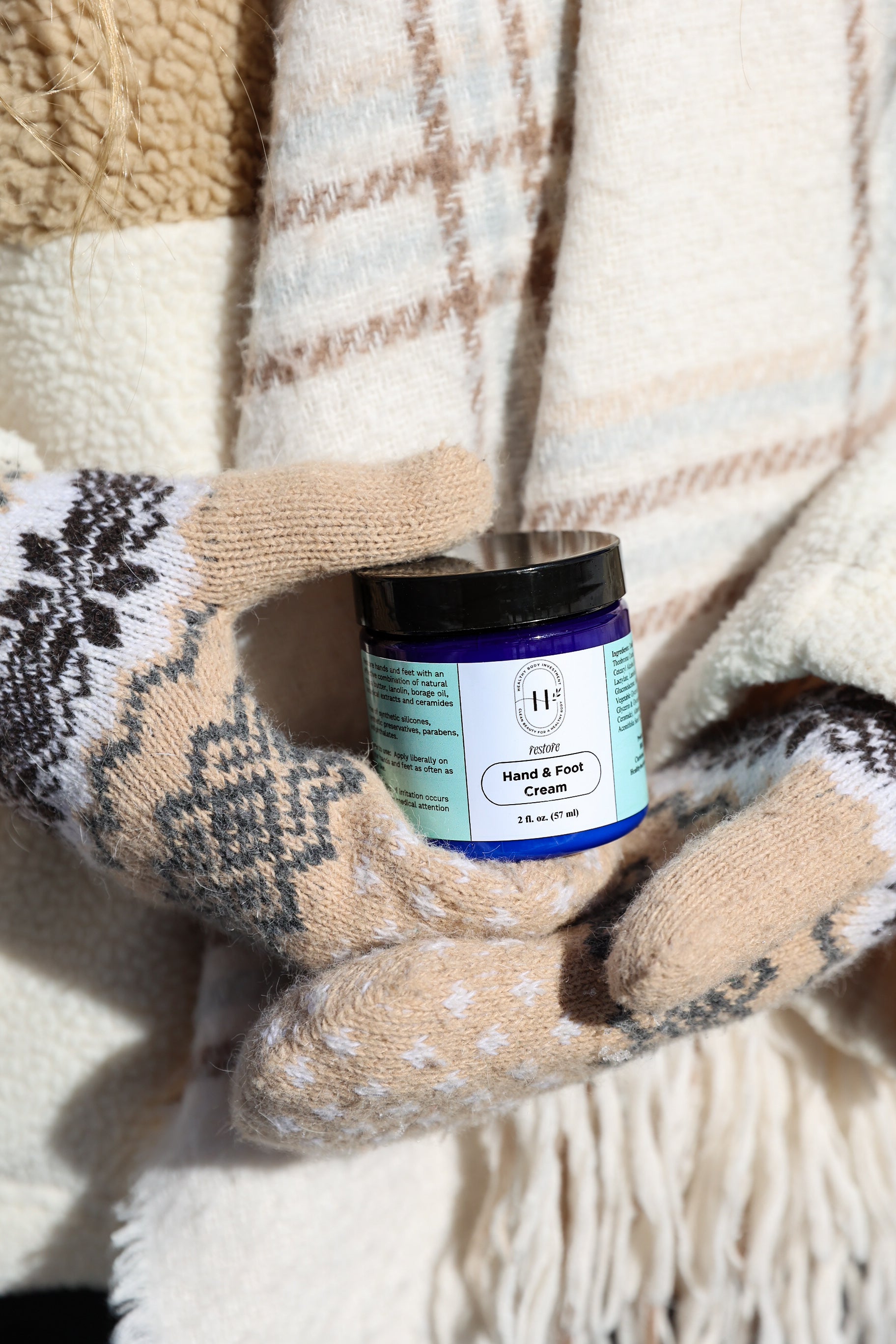 A jar of Restore Hand & Foot Cream held in a mitten-covered hand, with a plaid blanket in the background.