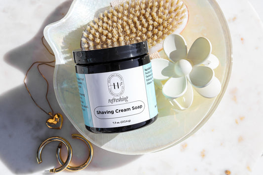 A jar of shaving cream soap labeled ' Inviting ' is displayed alongside a brush and a white flower on a light background.